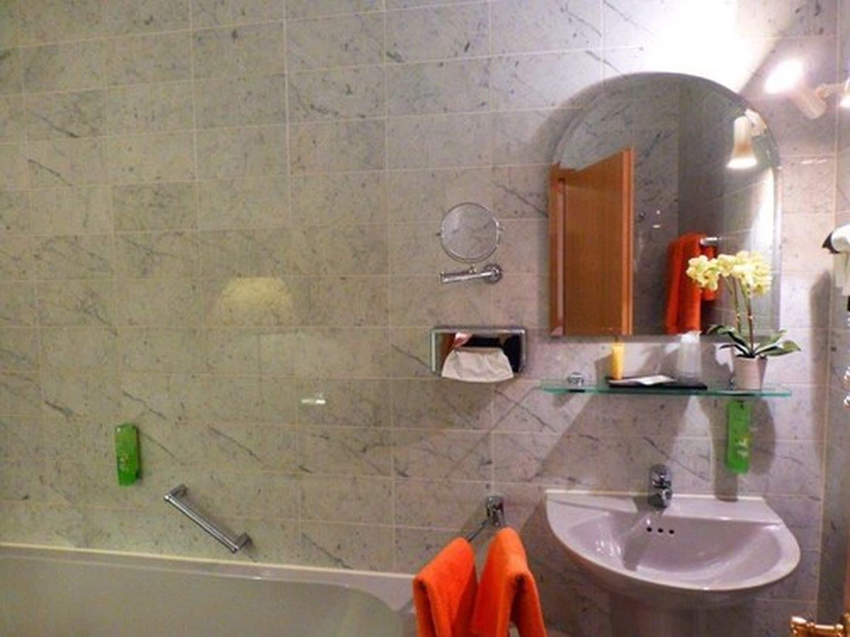 Ein Badezimmer mit einer Marmorwand, einem Waschbecken, einem Spiegel, einem Handtuchhalter mit orangefarbenen Handtüchern und einer Zimmerpflanze.