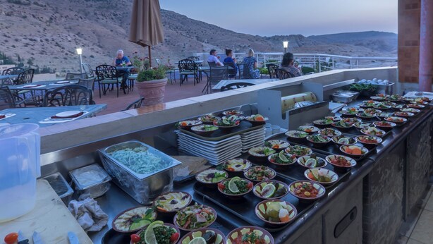 Un área de comedor al aire libre con varias mesas y sillas, un bufé con diversos platos, y vistas a un paisaje montañoso.