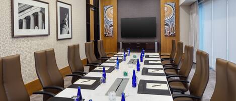 A conference room with a long table, brown leather chairs, a flat-screen TV, framed artwork, and a patterned wall.