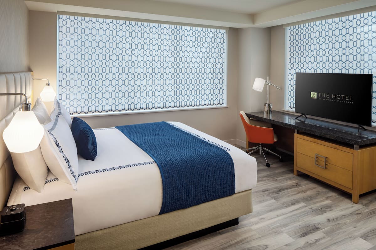 A hotel room with a bed, a desk with a television, a chair, and a nightstand with a lamp.