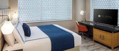 A hotel room with a bed, a desk with a television, a chair, and a nightstand with a lamp.