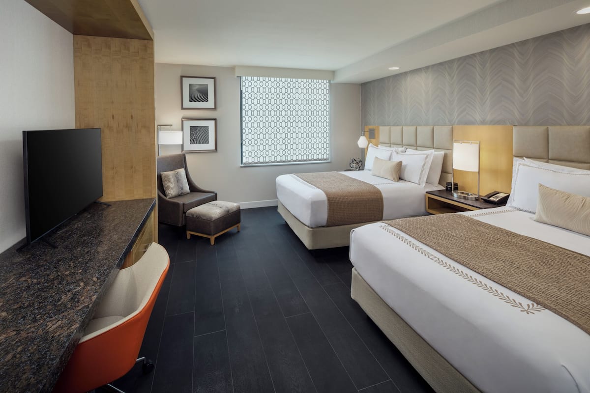A hotel room with two beds, a desk, a TV, and a window with a patterned blind.