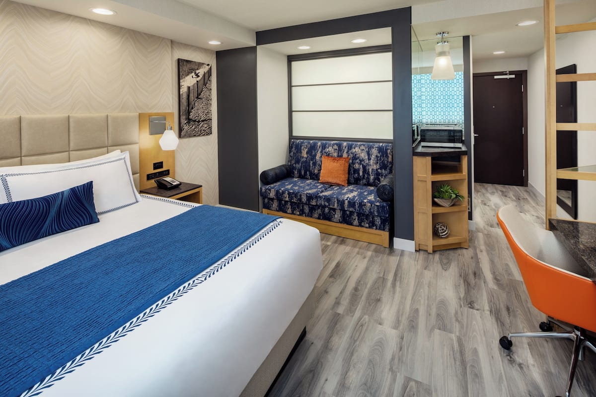A modern hotel room with a large bed, a sofa, a desk, and a small seating area.