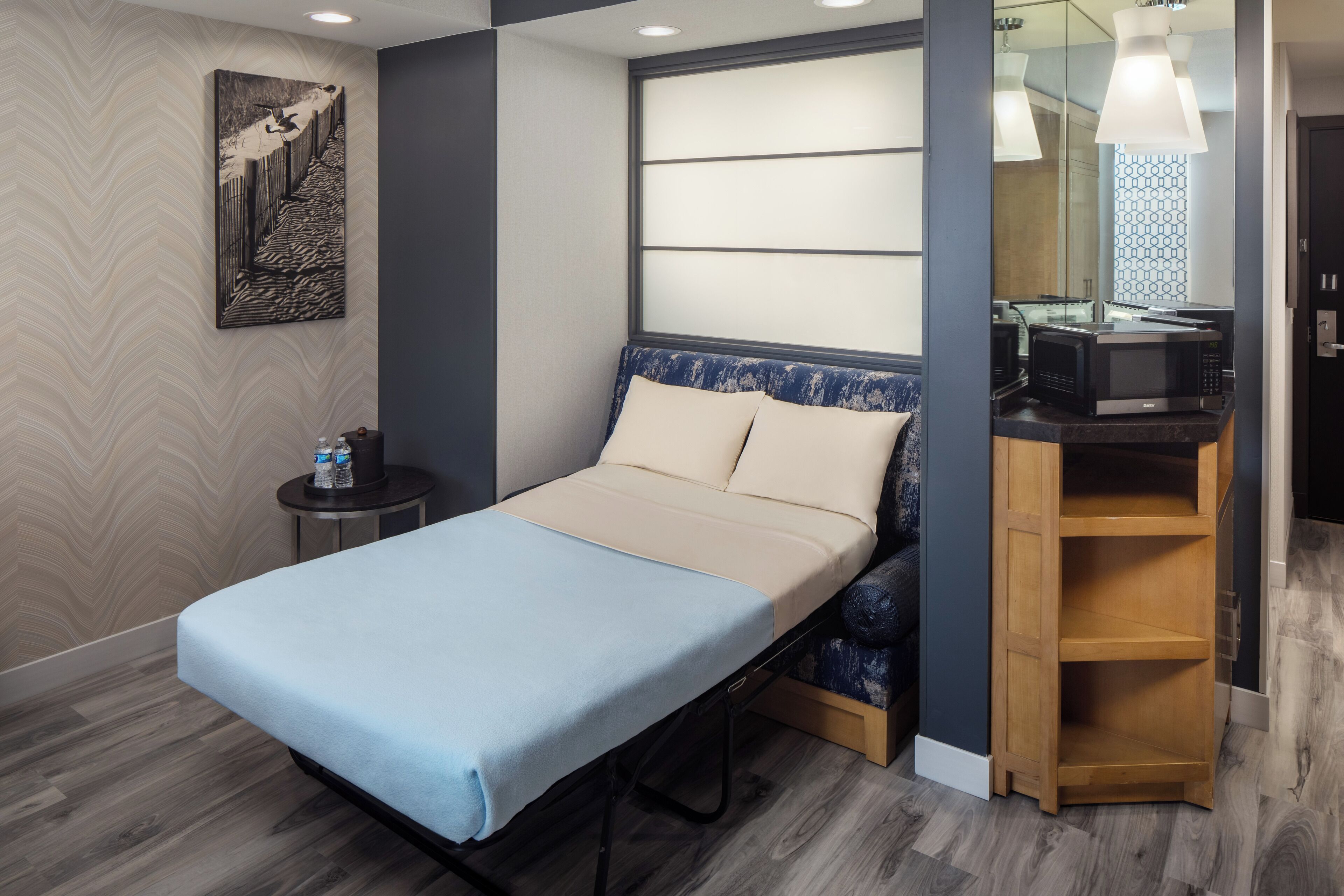 A compact bedroom with a single bed, a small bedside table, a microwave, and a wooden shelf unit.