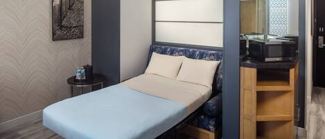 A compact bedroom with a single bed, a small bedside table, a microwave, and a wooden shelf unit.