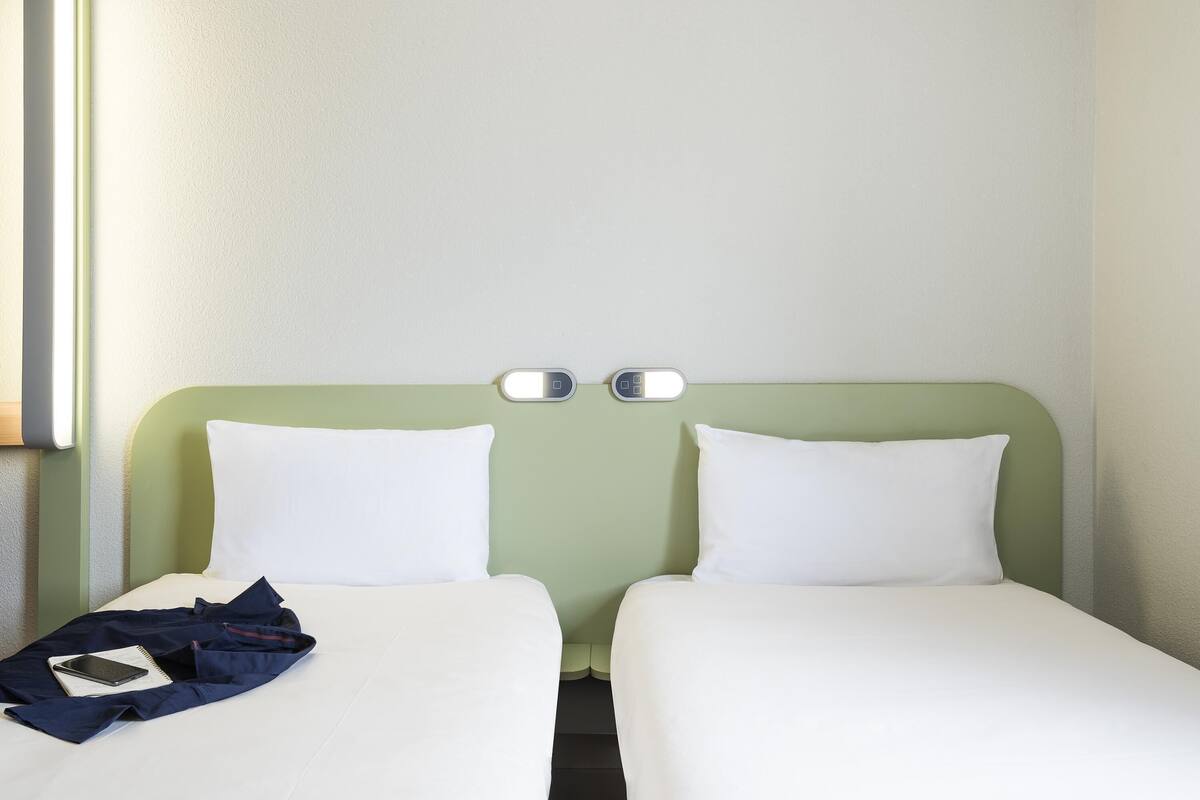 Quarto de hotel com duas camas de solteiro, mesa de cabeceira com abajur e luminária na parede.