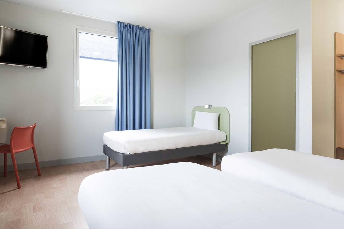 Quarto de hotel com cama, escrivaninha, cadeira, televisão e janela com cortinas.