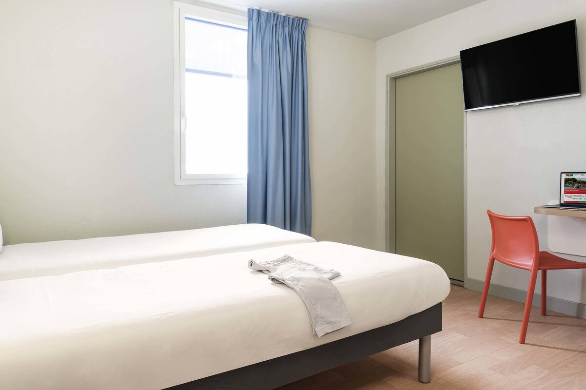 Quarto de hotel com uma cama, uma escrivaninha com cadeira, televisão e janela com cortinas.