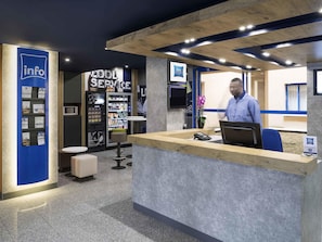 Una recepción con un hombre detrás, una máquina expendedora y un mostrador informativo azul.