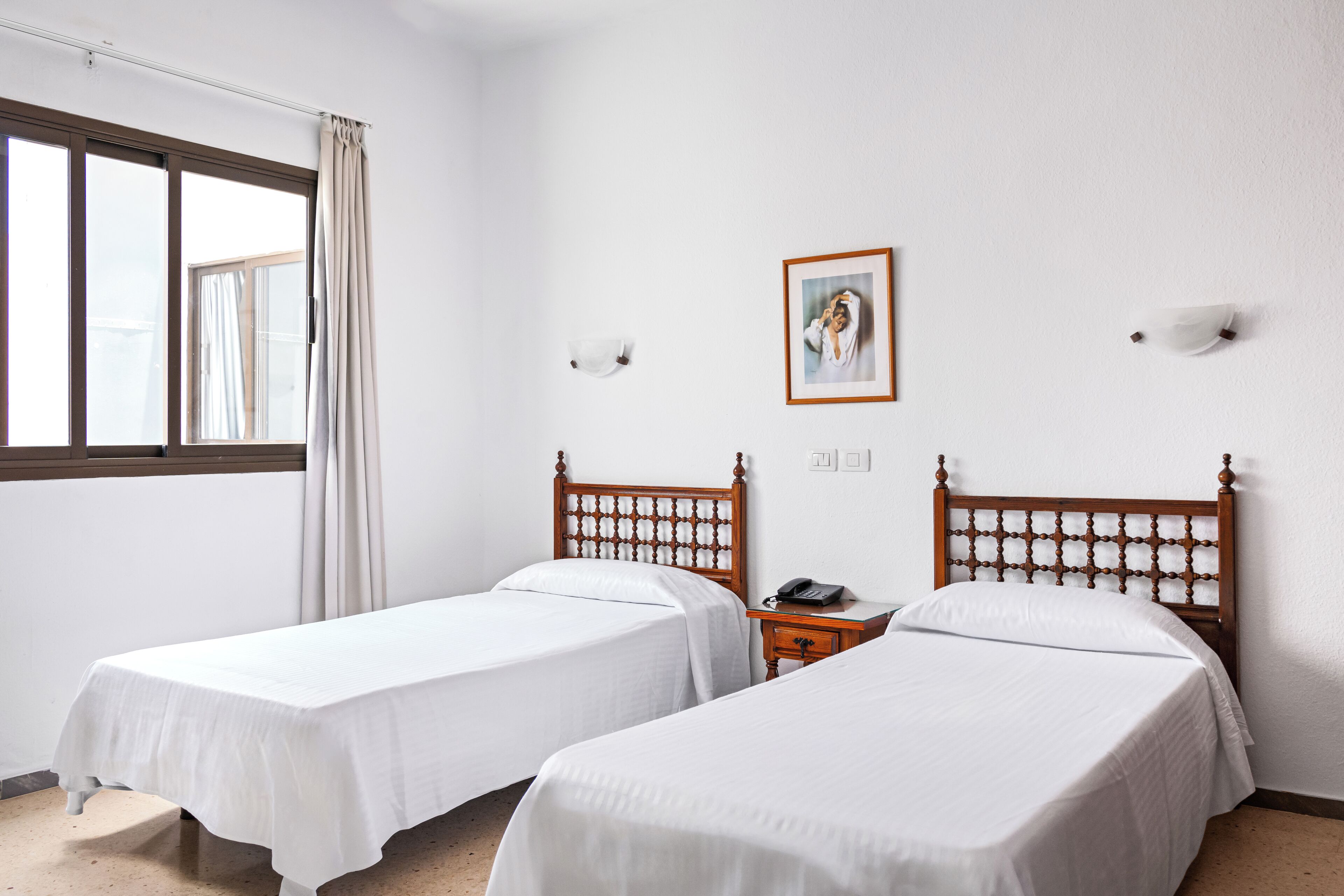 A hotel room with two single beds, a bedside table with a phone, a framed picture on the wall, and a window with curtains.