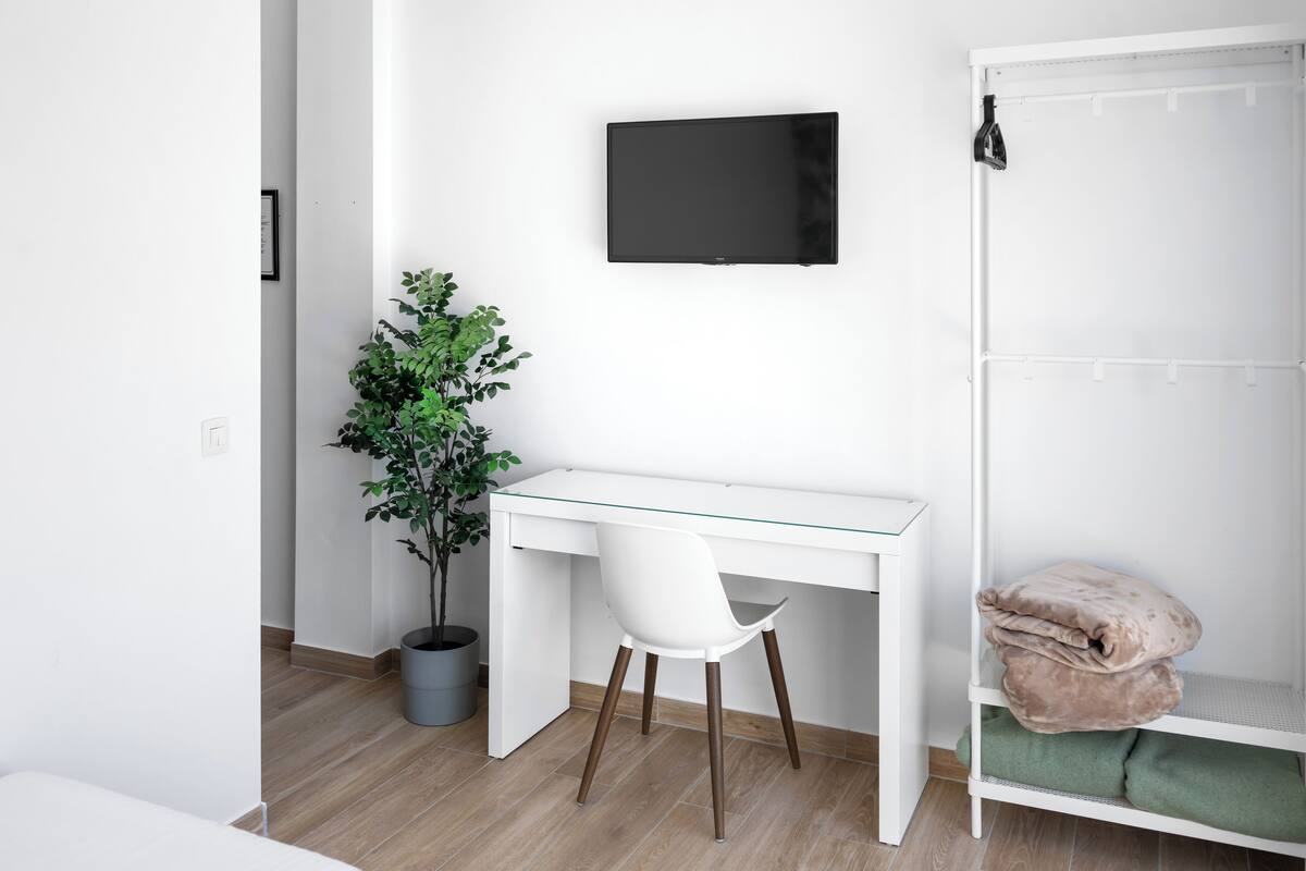 Habitación moderna con escritorio blanco, televisión montada, planta en maceta y cama con manta verde.