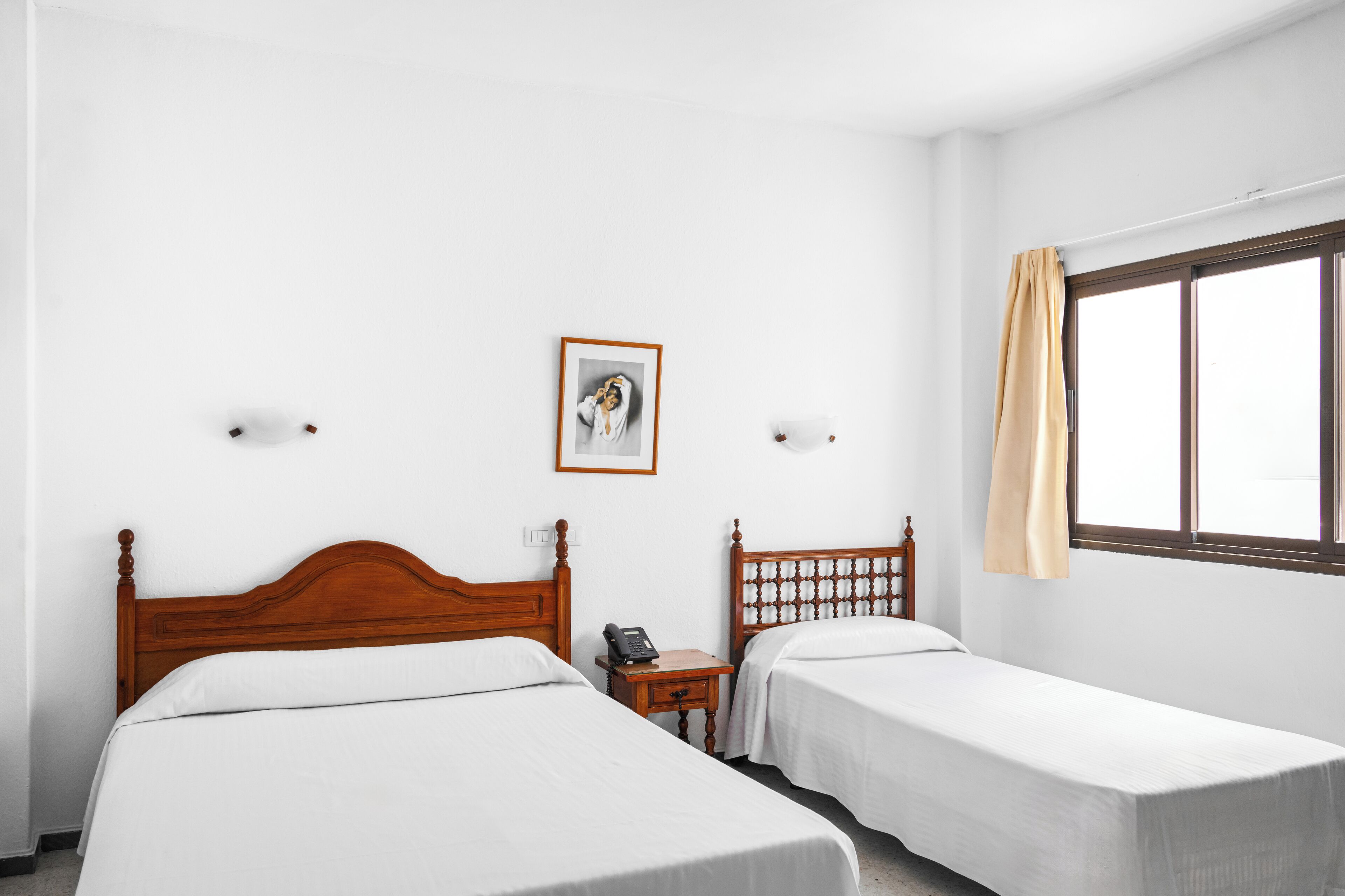 Een hotelkamer met twee eenpersoonsbedden, een houten hoofdbord, een nachtkastje met een telefoon, een ingelijst schilderij aan de muur en een raam met gordijn.