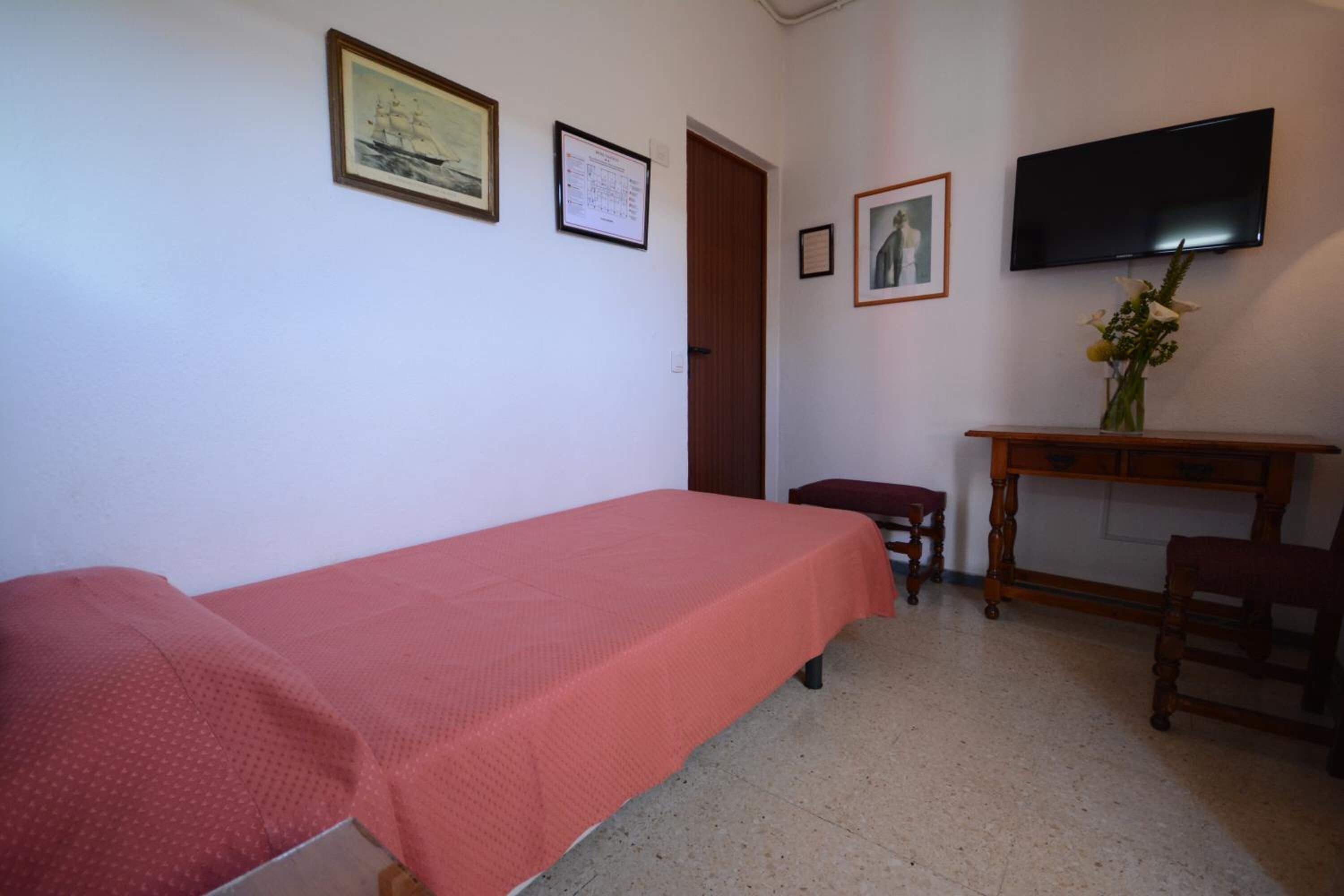 Une chambre avec un lit simple, un bureau en bois, une chaise, une télévision et des tableaux encadrés accrochés au mur.