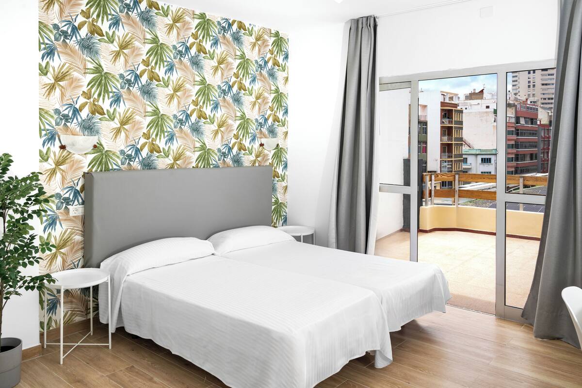 Una habitación de hotel moderna con una cama grande, una mesita pequeña, una planta en maceta y un balcón con vistas a la ciudad.