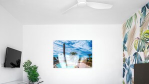 Une chambre moderne avec un ventilateur de plafond blanc, un téléviseur fixé au mur, un grand mur décoré sur le thème des tropiques et un tableau représentant une piscine et des palmiers.