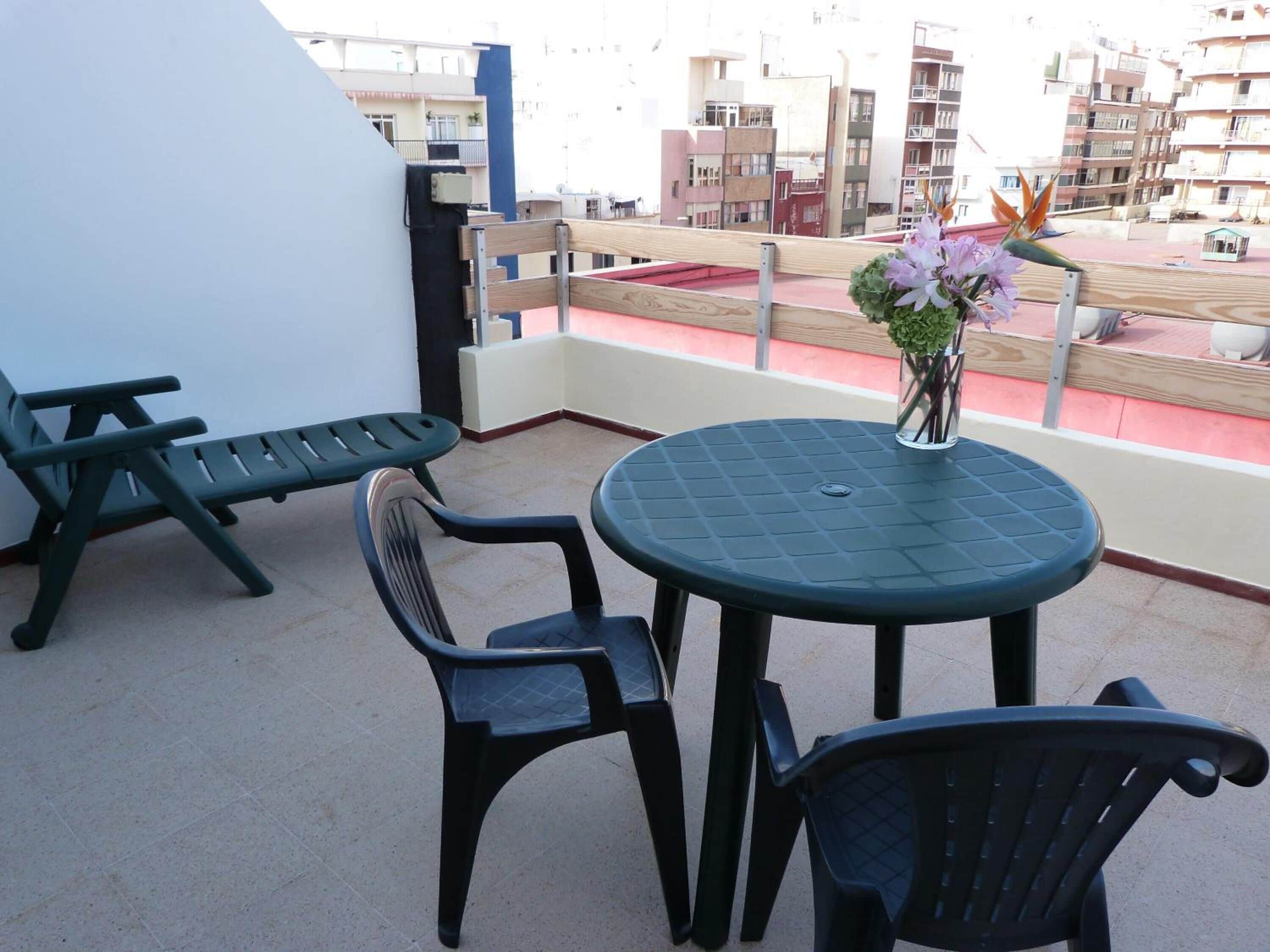Un balcón con una mesa redonda y dos sillas, un jarrón pequeño con flores y vista a edificios al fondo.