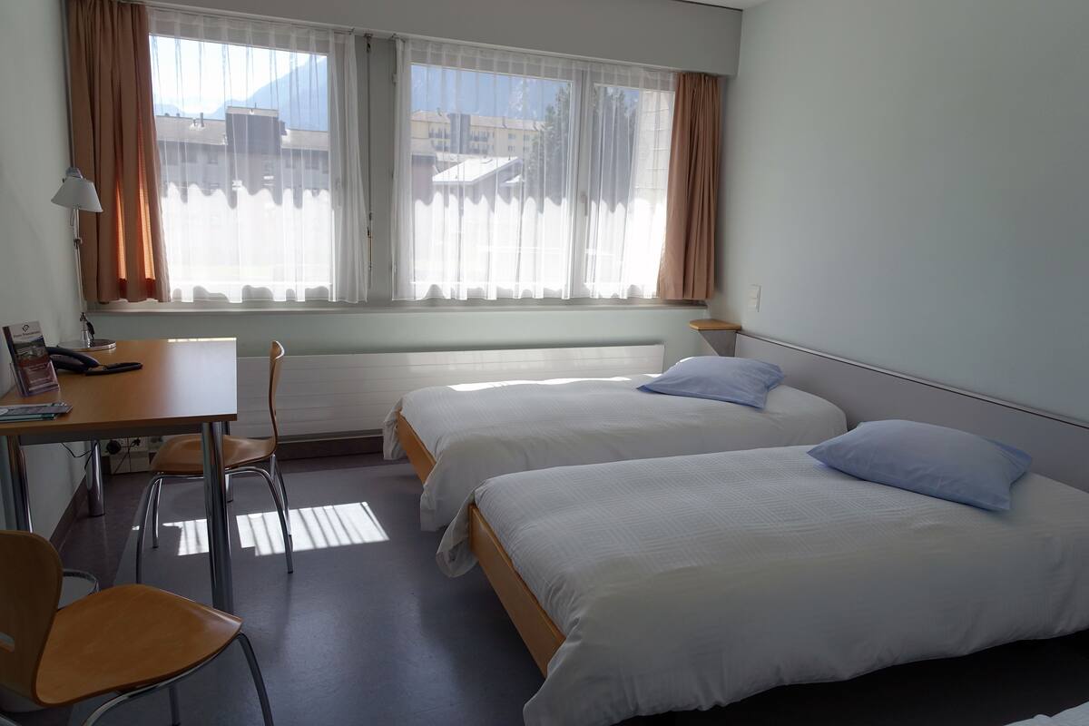 Une chambre d’hôtel avec deux lits simples, un bureau avec une chaise et une fenêtre avec des rideaux.