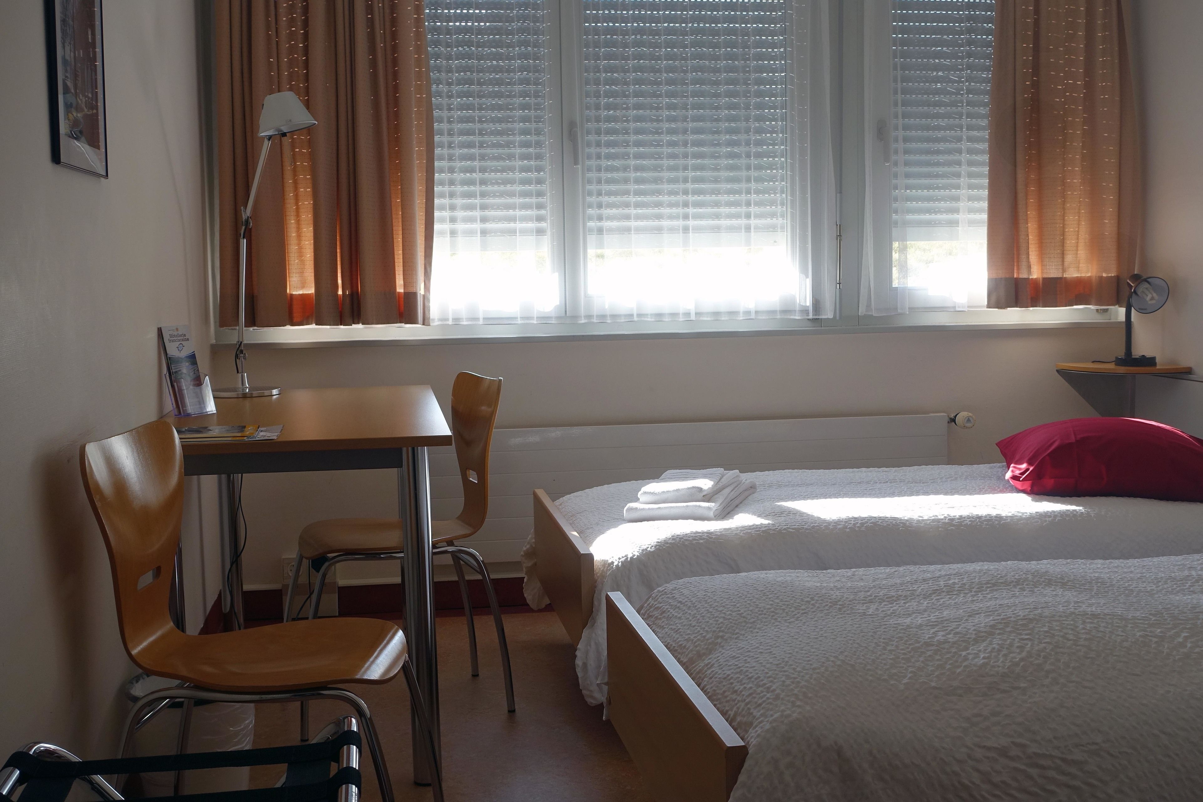 A hotel room with two beds, a desk, a chair, a lamp, and a window with curtains.