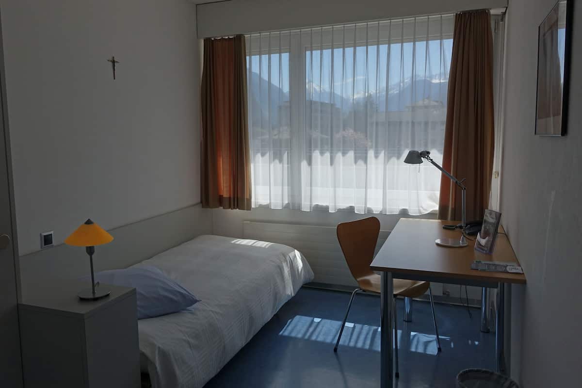 Une chambre d’hôtel avec un lit, un bureau et une chaise, une lampe et une fenêtre avec des rideaux.