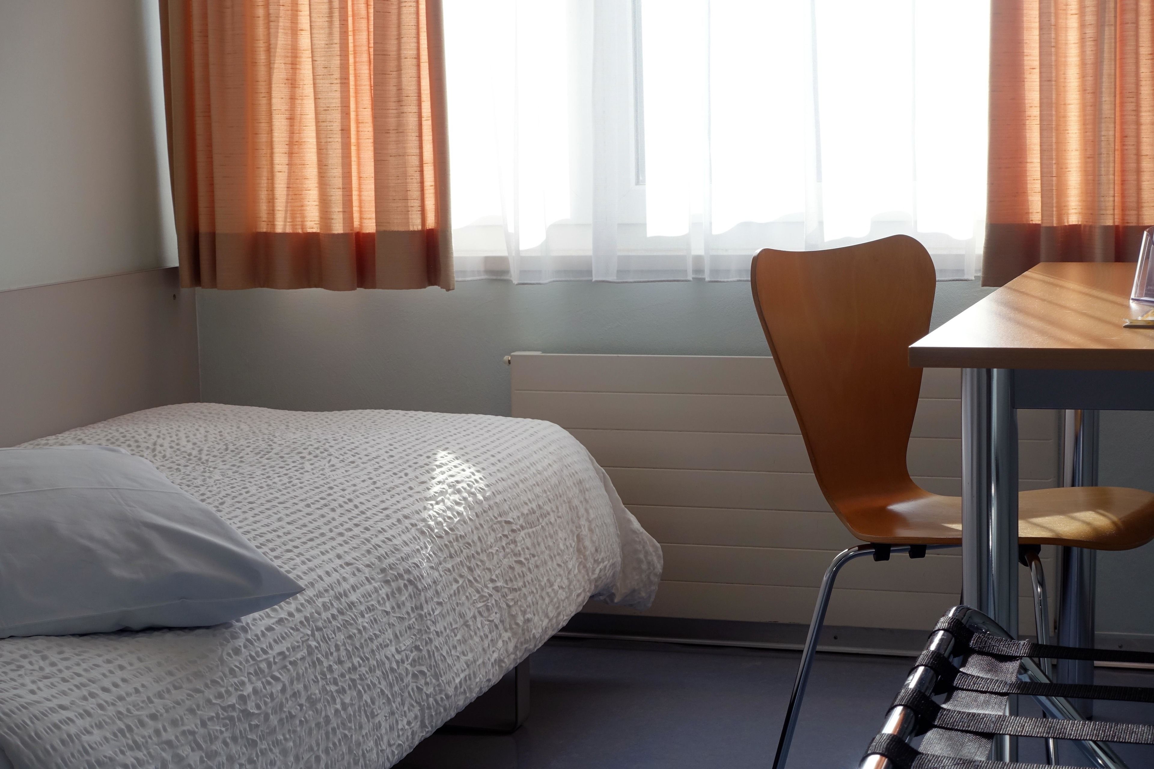 Una habitación de hotel con una cama individual, una silla de madera, una mesita pequeña y una ventana con cortinas de gasa.