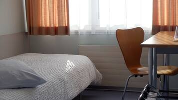 Una habitación de hotel con una cama individual, una silla de madera, una mesita pequeña y una ventana con cortinas de gasa.