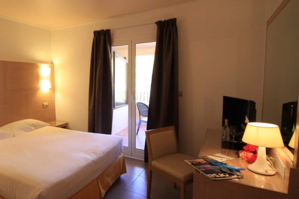 A hotel room with a bed, bedside tables, a desk with a lamp, and a view through a door.