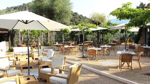 An outdoor seating area with wicker furniture and umbrellas, surrounded by trees and hills.
