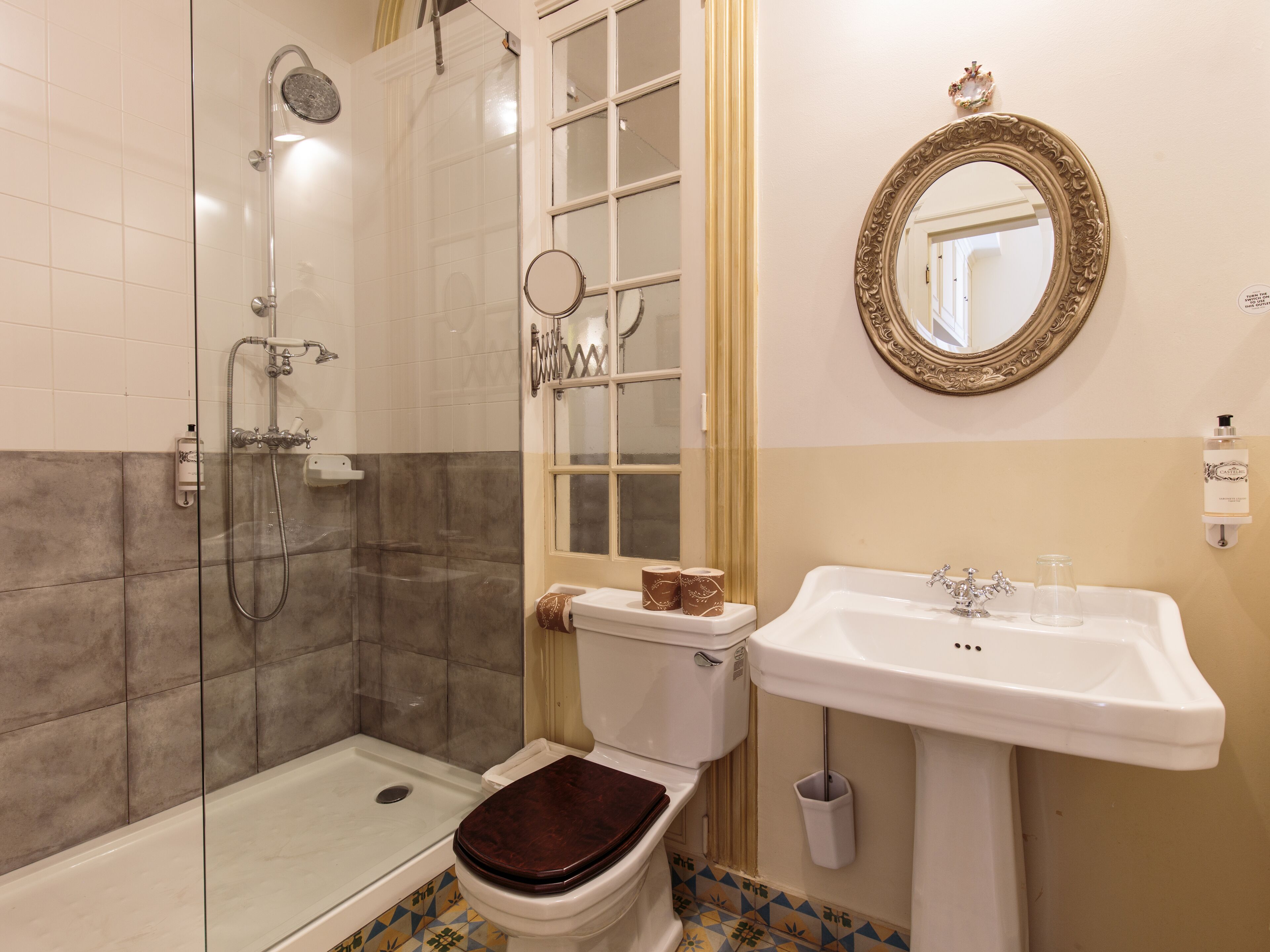 A bathroom with a glass-enclosed shower, a toilet with a brown seat, a sink with a faucet, and a mirror with an ornate frame.