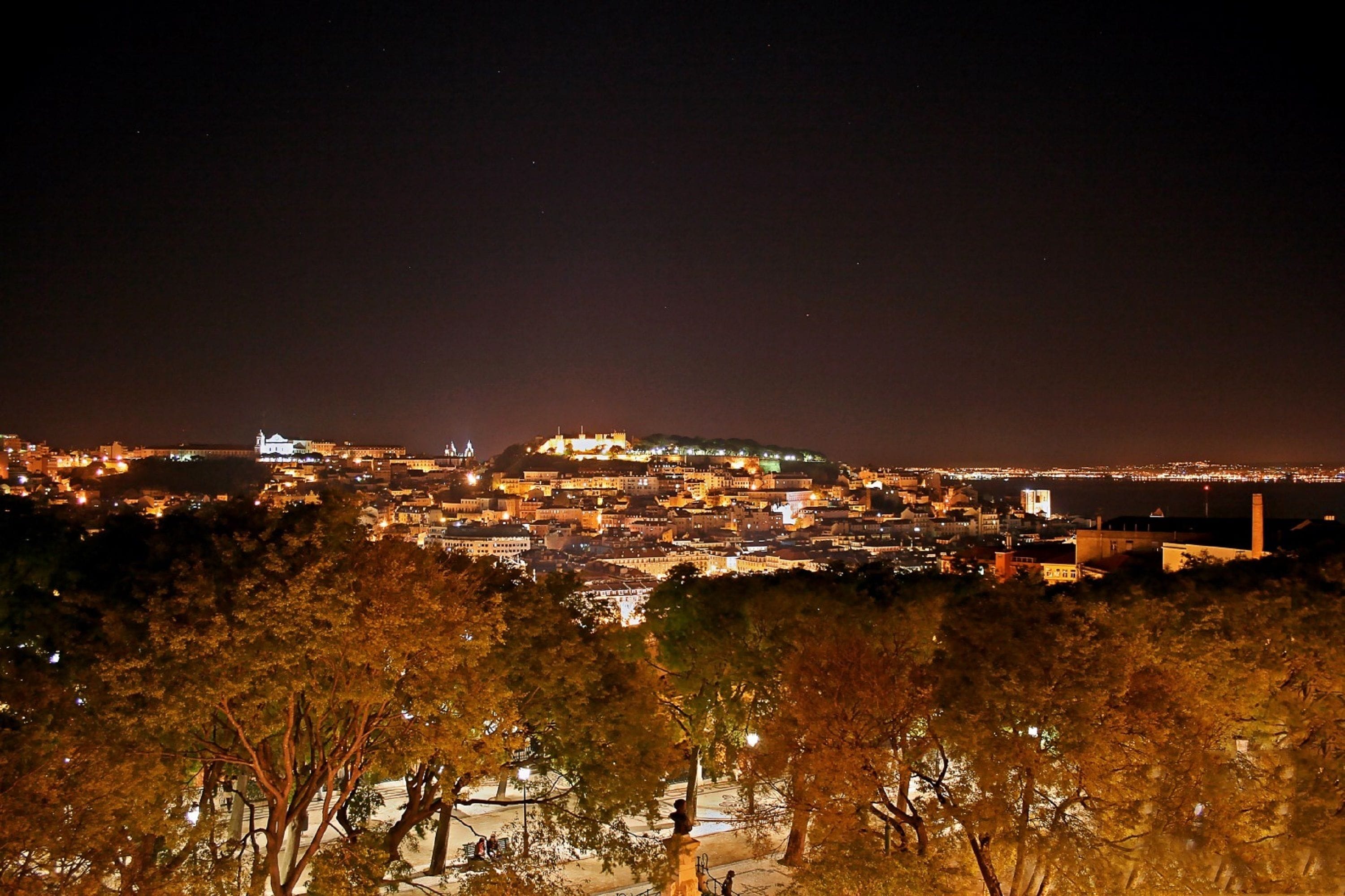 A nighttime cityscape with a prominent hilltop building, illuminated streets, and a distant view of the coastline.