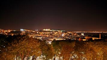 Una vista nocturna de la ciudad con un edificio destacado en la cima de una colina, calles iluminadas y una costa visible a lo lejos.