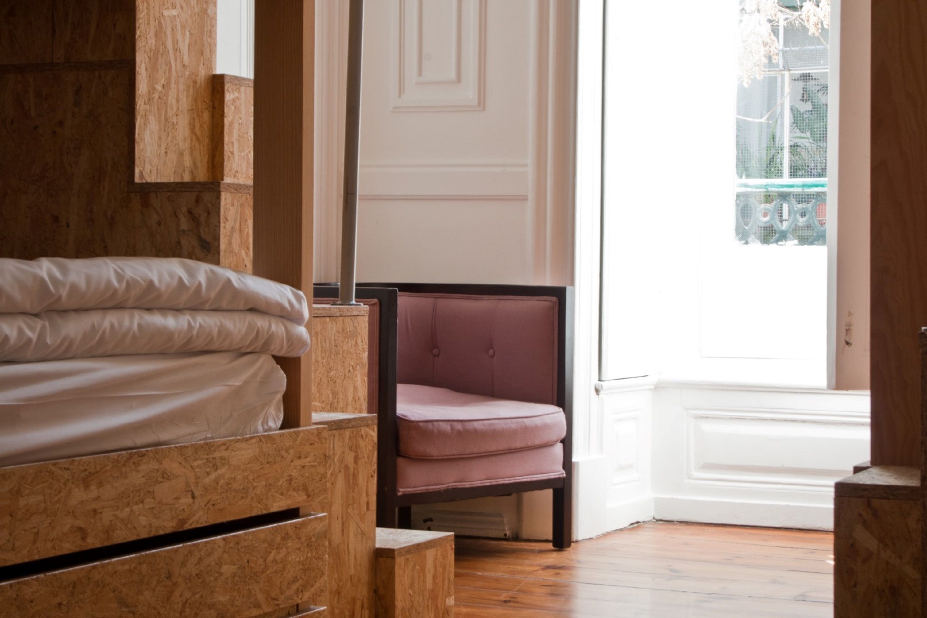A bedroom with a wooden bed, a pink chair, and a window with a view of greenery.