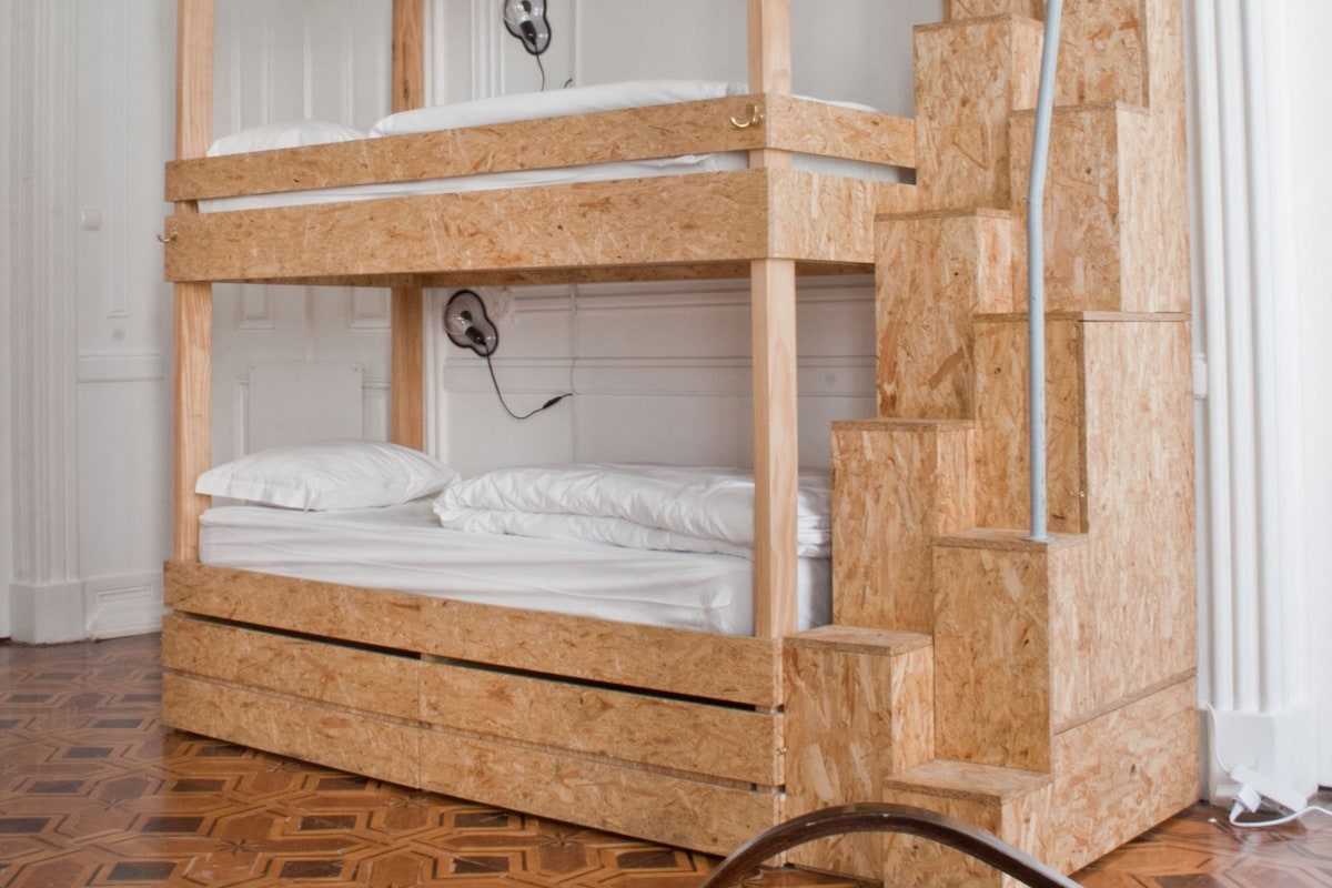 A bunk bed with a wooden frame and white bedding, located in a room with patterned flooring and white walls.