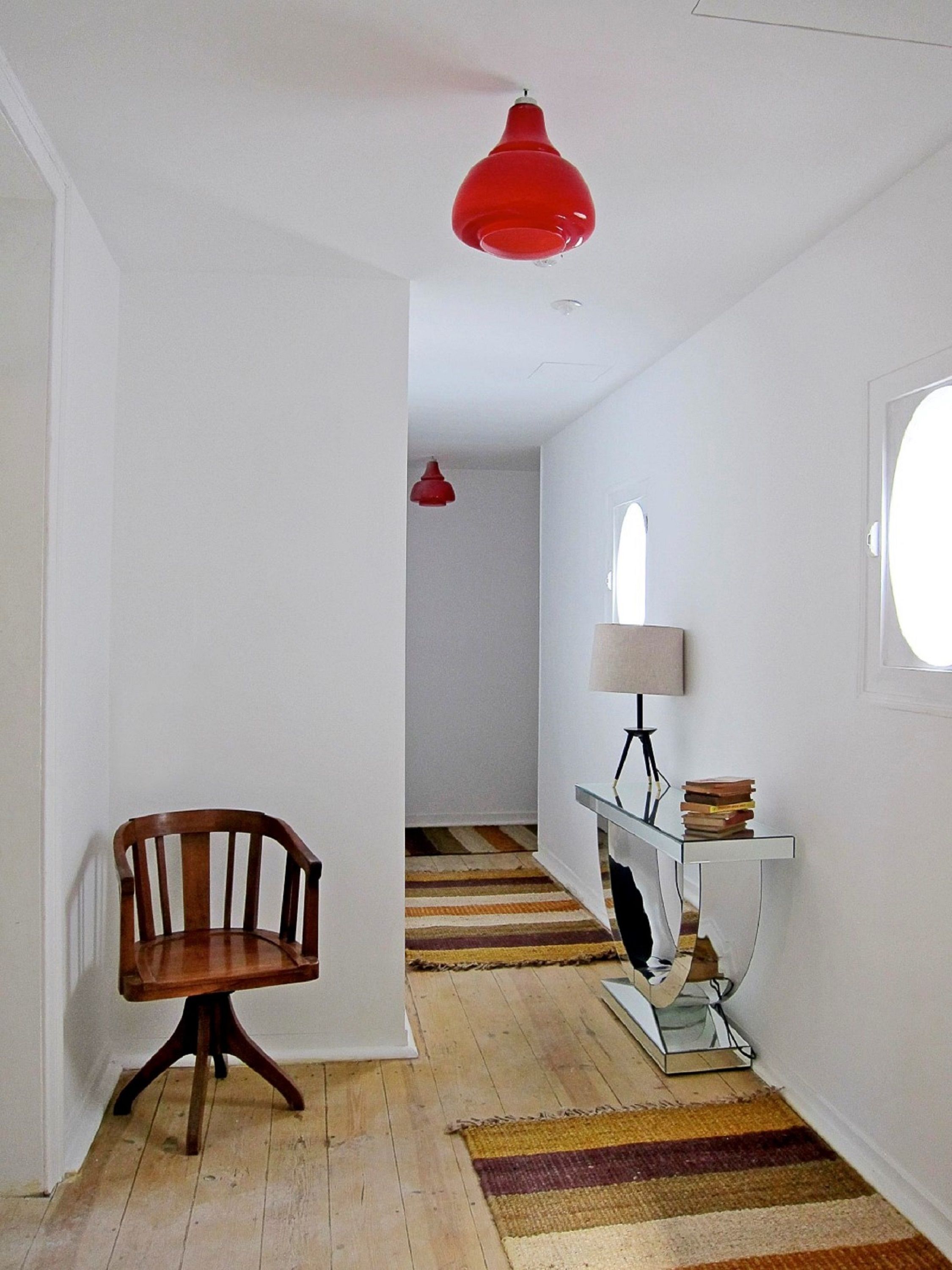 Un pasillo con una silla de madera, una mesita de cristal con libros, una lámpara colgante roja y una ventana a la derecha.