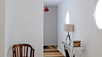 Un pasillo con una silla de madera, una mesita de cristal con libros, una lámpara colgante roja y una ventana a la derecha.