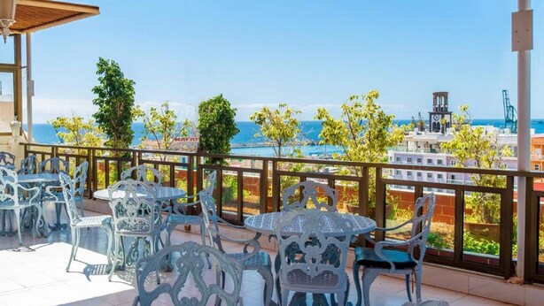 Un patio exterior con sillas y mesas de metal ornamentado, con vistas a un paisaje urbano costero y una vista despejada del mar y un barco a lo lejos.