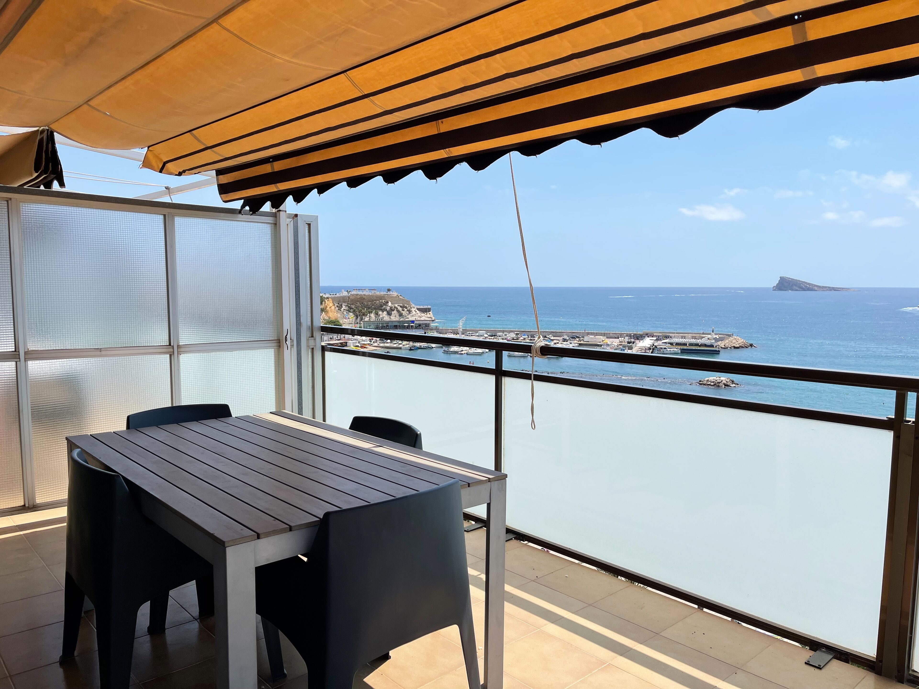 Un balcón con mesa y sillas, con vista a una zona costera con barcos y un promontorio rocoso.