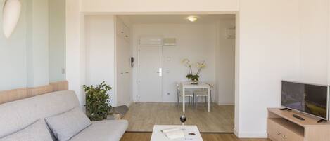 Una sala de estar moderna con un sofá, una mesa de centro con un libro y un vaso, un televisor sobre un mueble de madera y una zona de comedor al fondo.