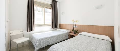 A bedroom with two single beds, a chair, a small table with a plant, and a window with curtains.