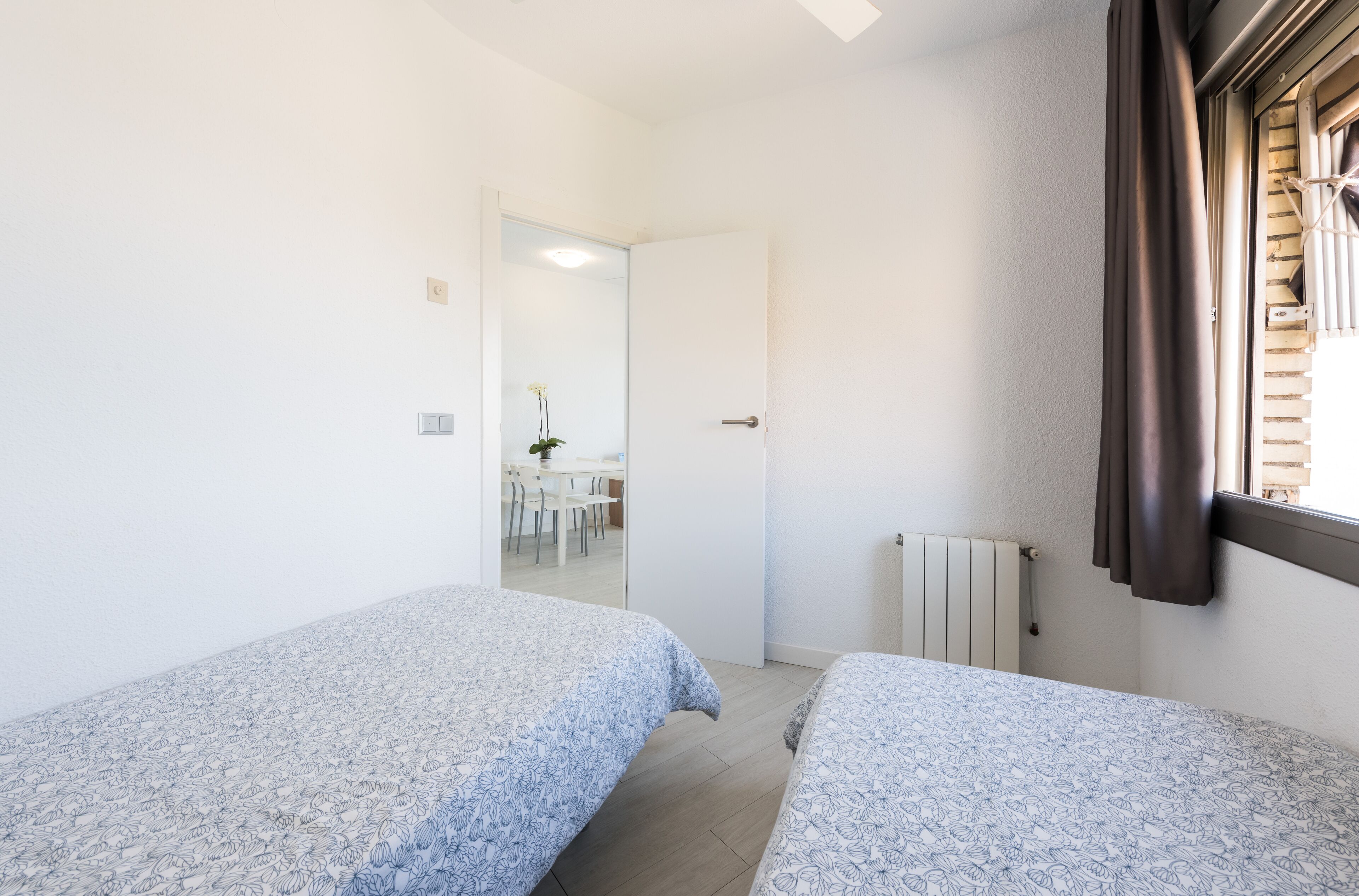A bedroom with two beds, a window with blinds, a radiator, and a door leading to a dining area.