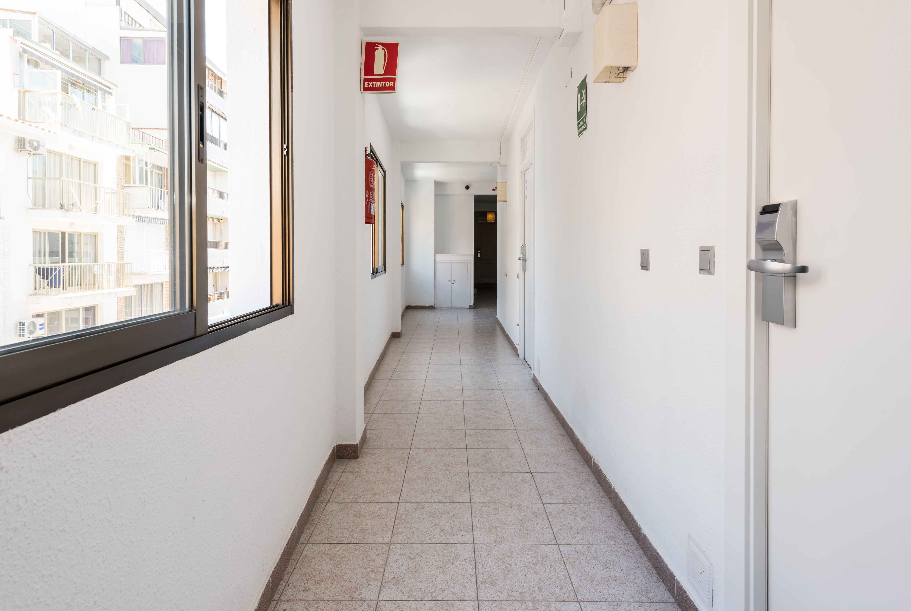 A corridor with white walls, a tiled floor, and a fire exit sign.