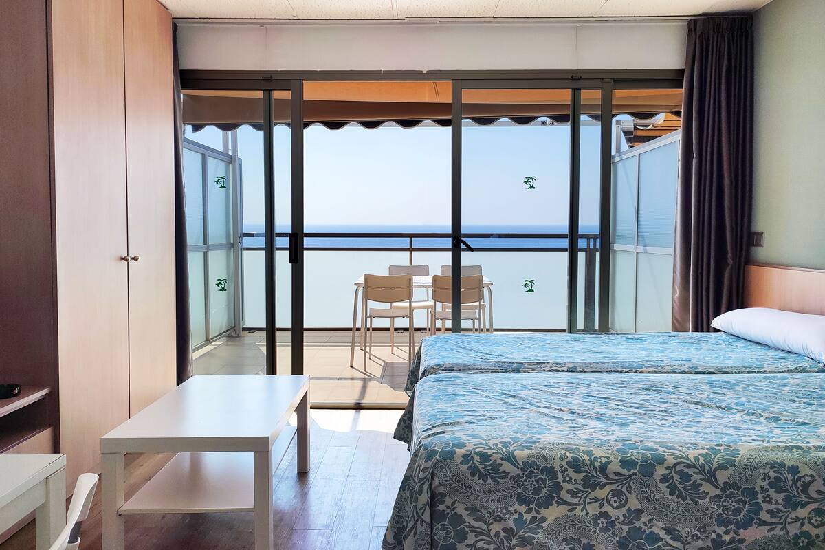 A hotel room with a balcony, a bed with a patterned bedspread, a small table, and two chairs.