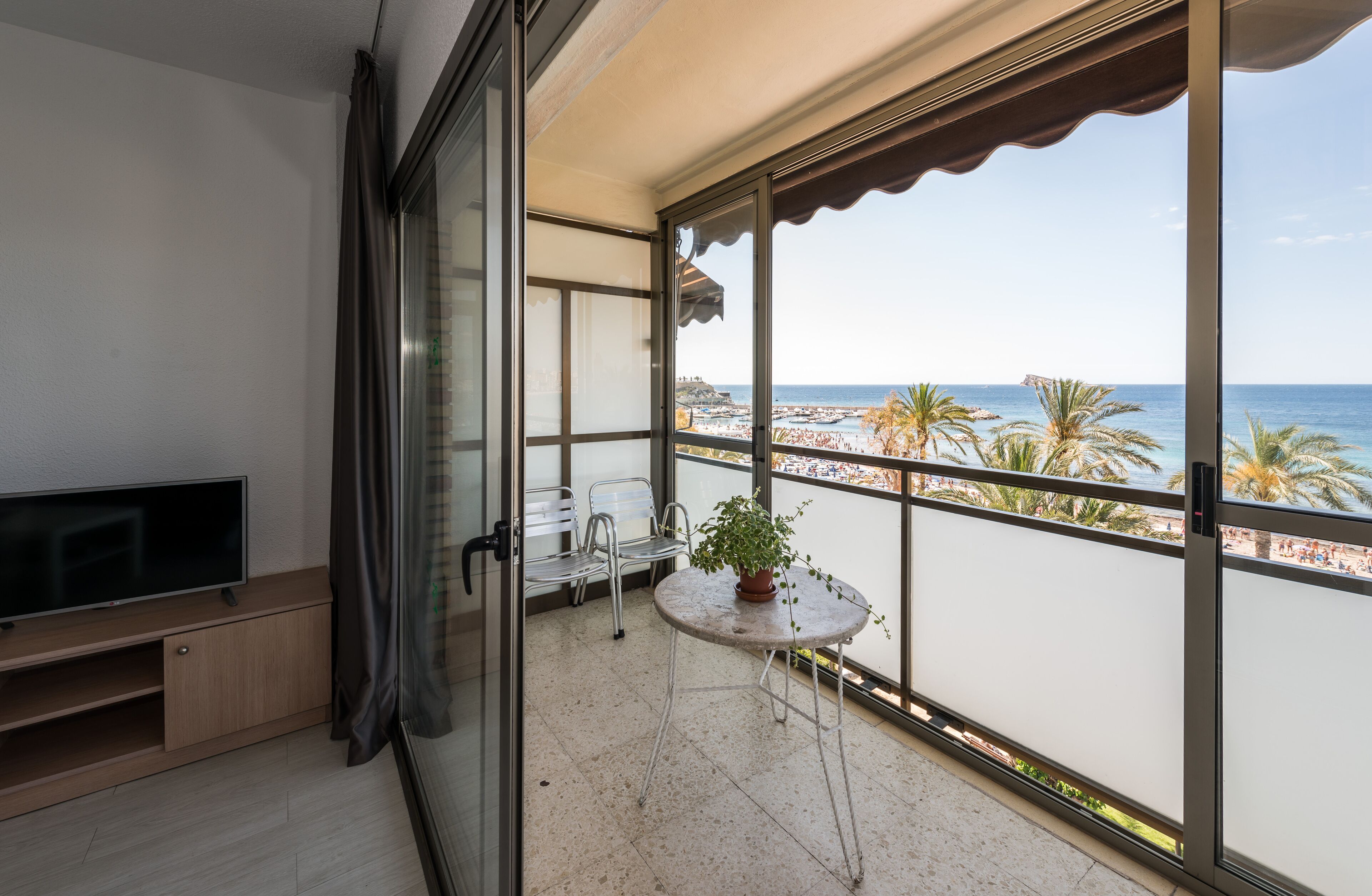 Ein Balkon mit Fernseher, Sitzgelegenheit und Topfpflanze mit Blick auf Strand und Meer.