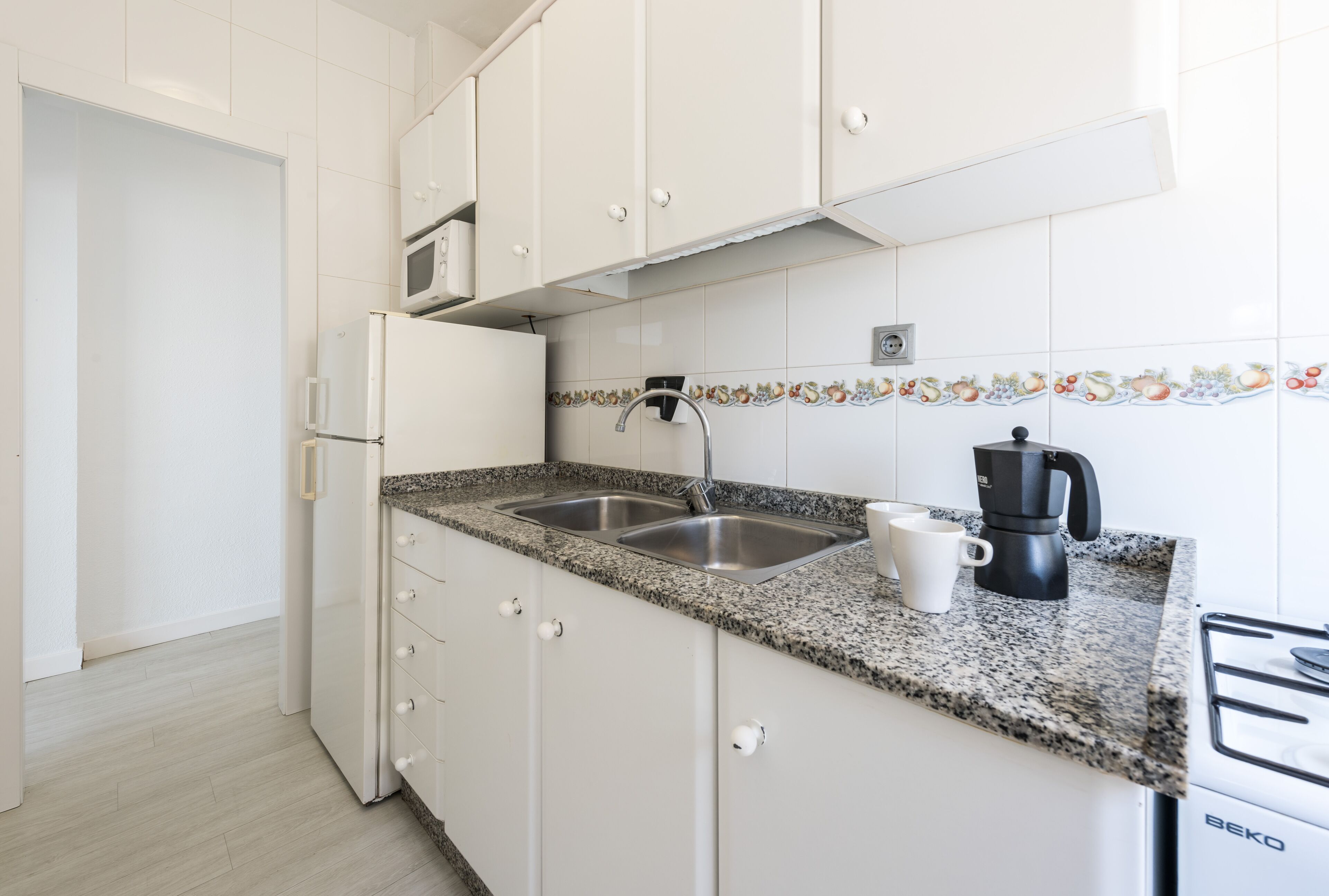 Una cucina con mobili bianchi, un piano di lavoro in granito, un lavello a due vasche, un forno a microonde, una macchina da caffè e un forno Beko.