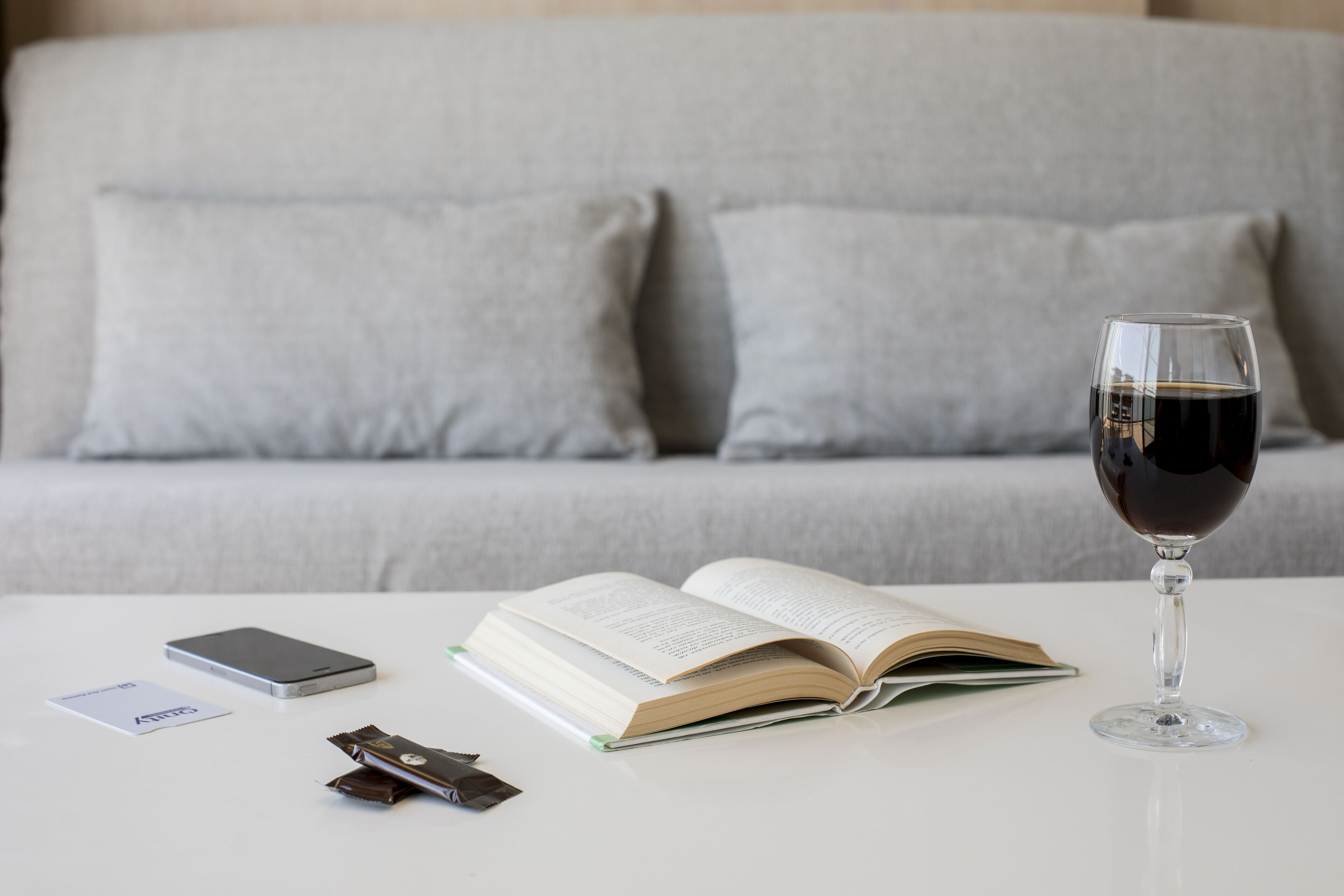 Une table blanche avec un livre ouvert, un verre de vin rouge, un téléphone intelligent et deux barres de chocolat.