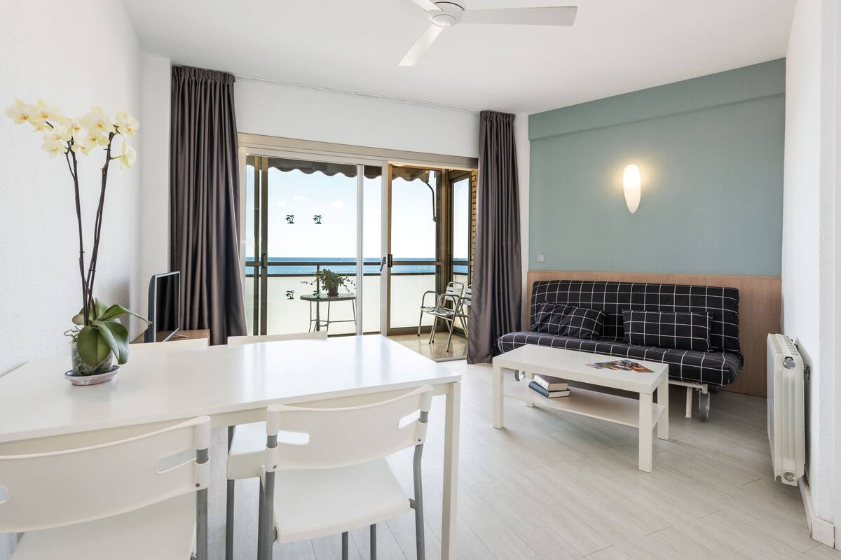 A modern living room with a white table, a black and white checkered sofa, and a view of the sea through large glass doors.