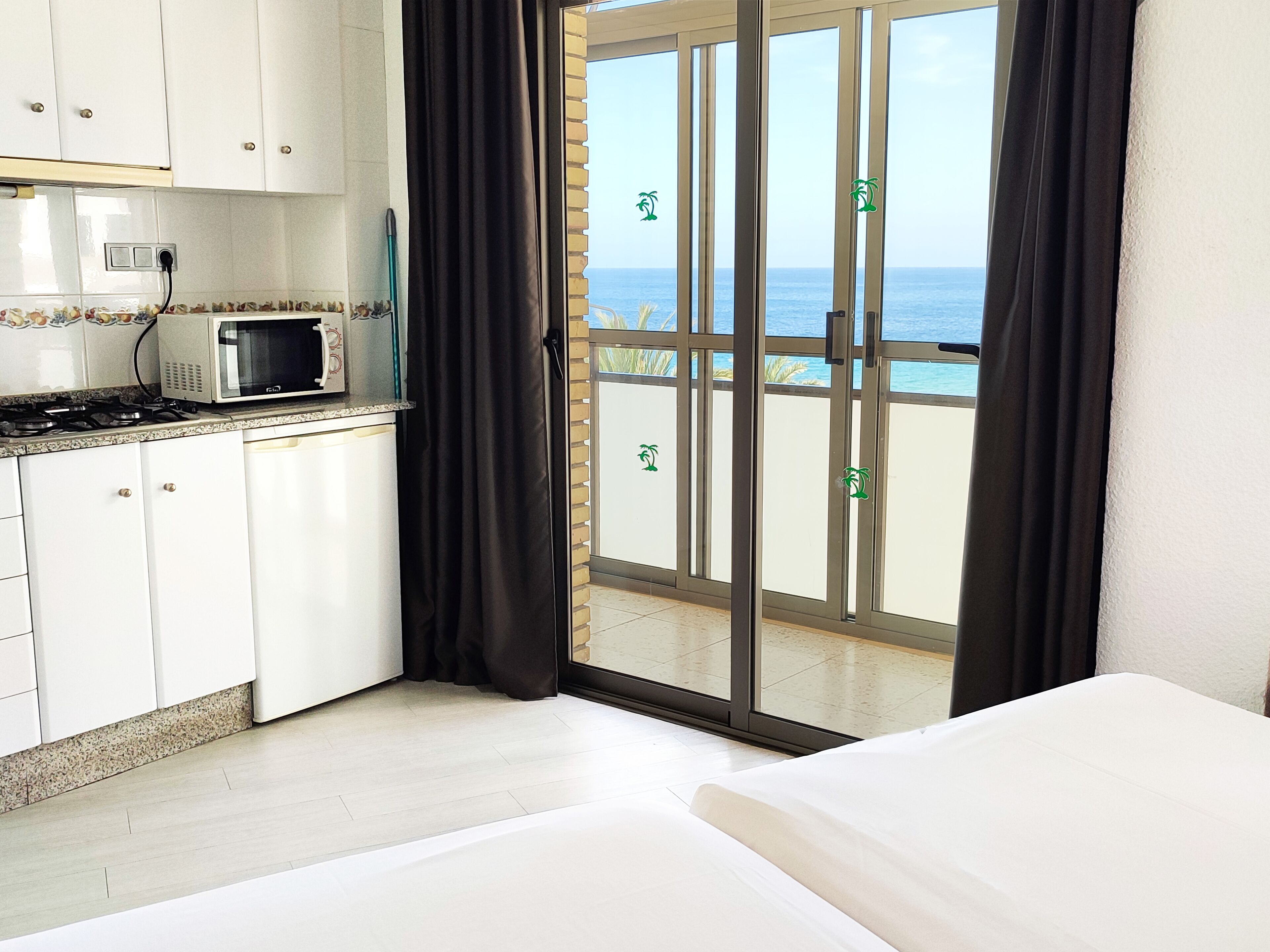 A room with a kitchenette, a bed, and a sliding glass door leading to a balcony with a view of the sea.