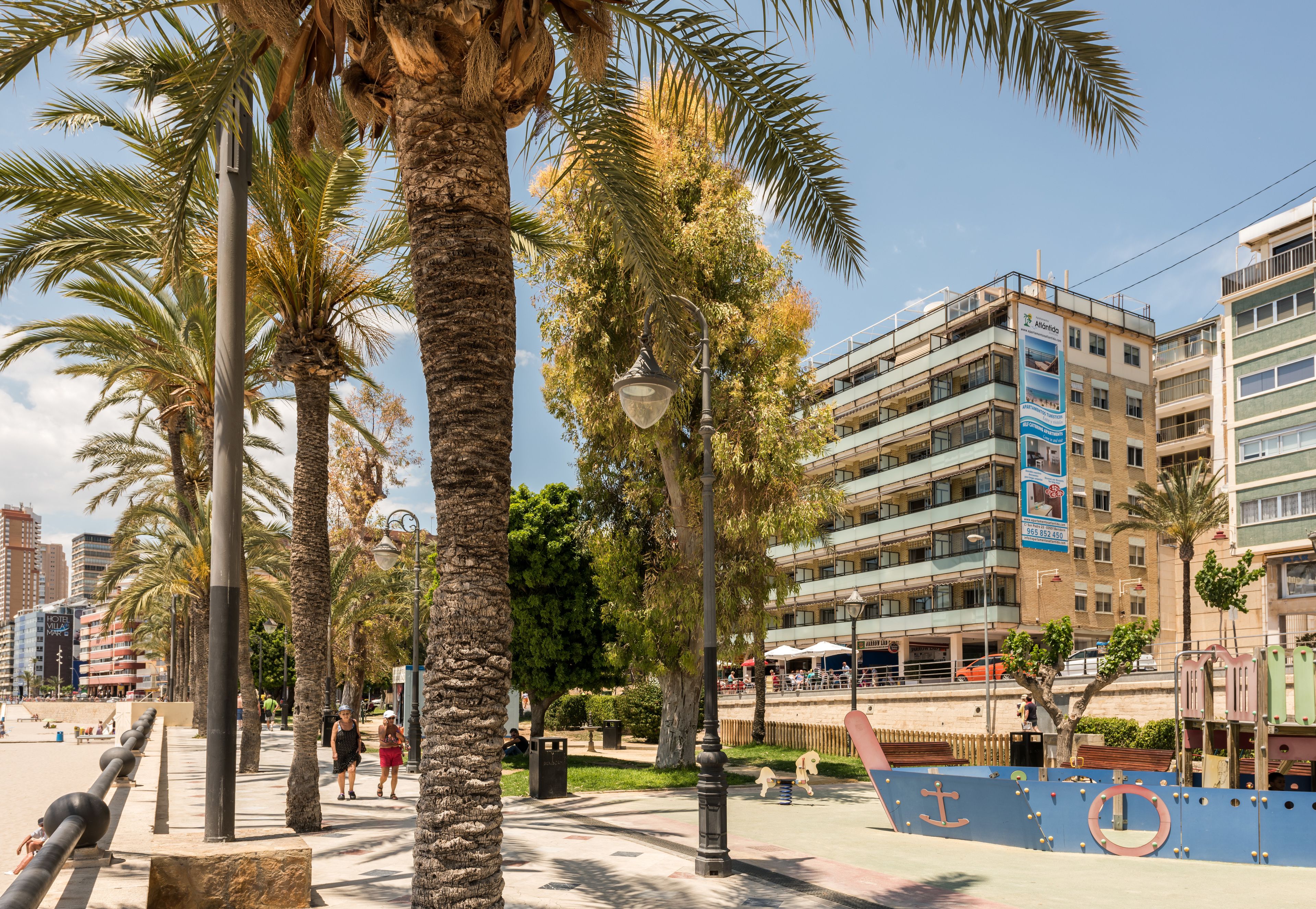 A promenade with palm trees, a blue mural, and a multi-story building with balconies.