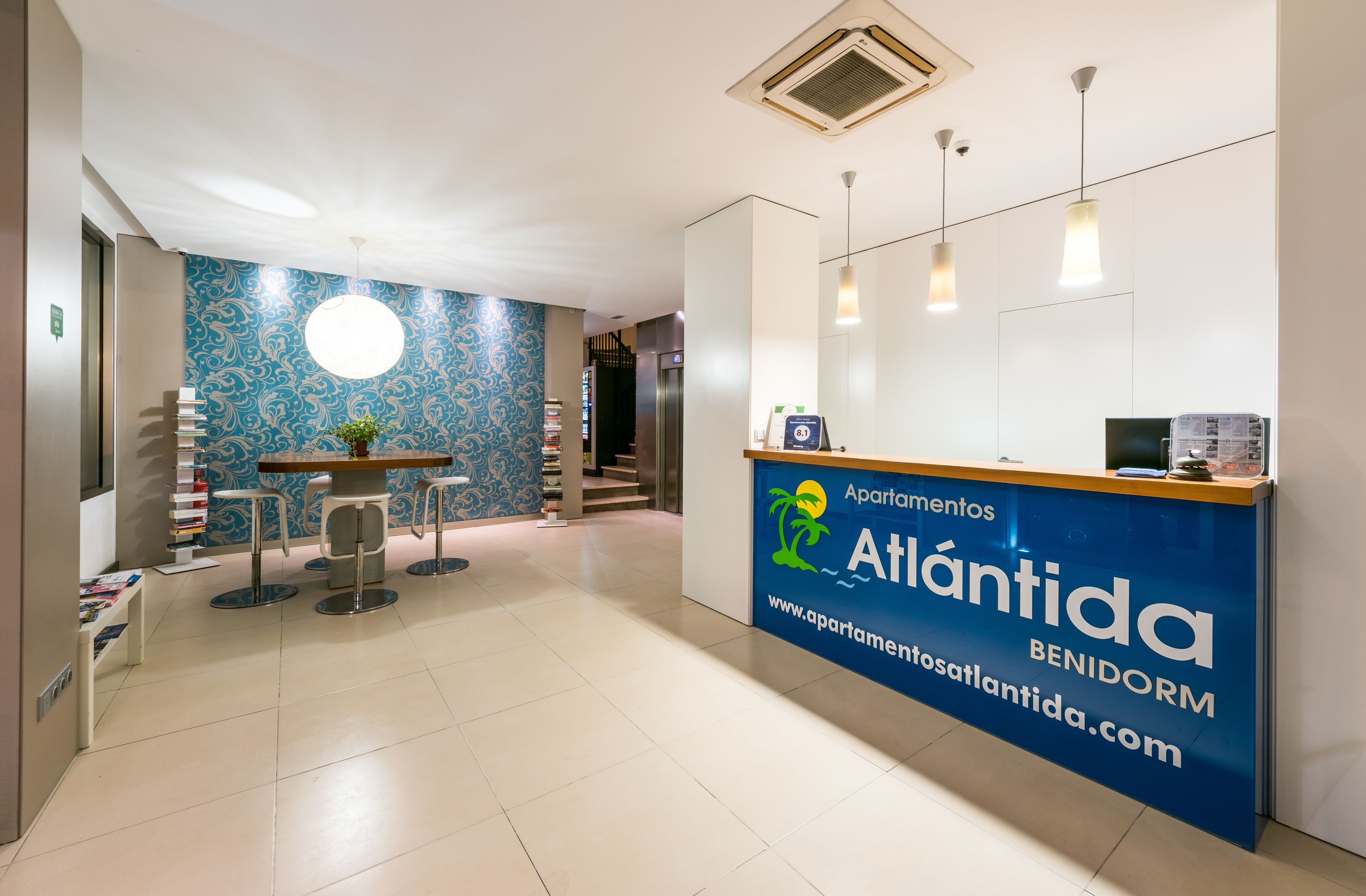 Een receptiegedeelte met een blauwe balie waarop de naam 'Atlantida' staat en een website-URL. Er zijn twee barkrukken en een ronde tafel met een plant. De vloer is betegeld, en de muren hebben een blauw patroon.