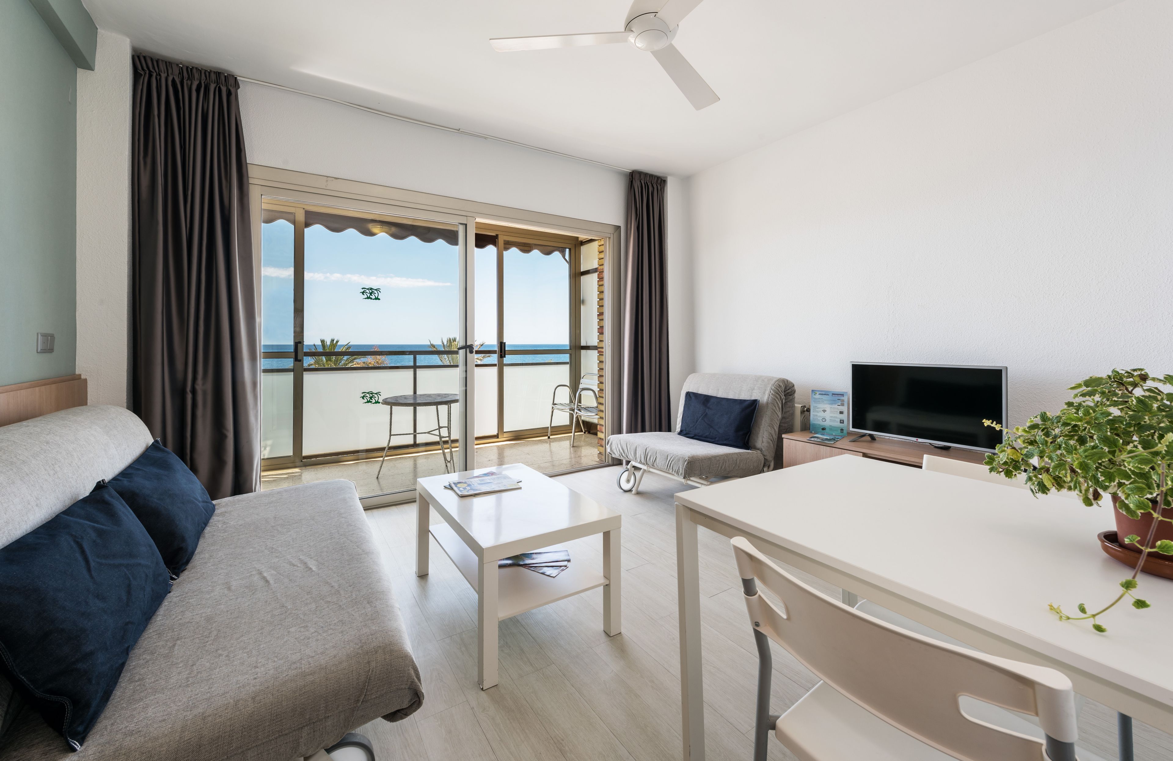 A modern hotel room with a balcony, a sofa, a dining table, and a TV.