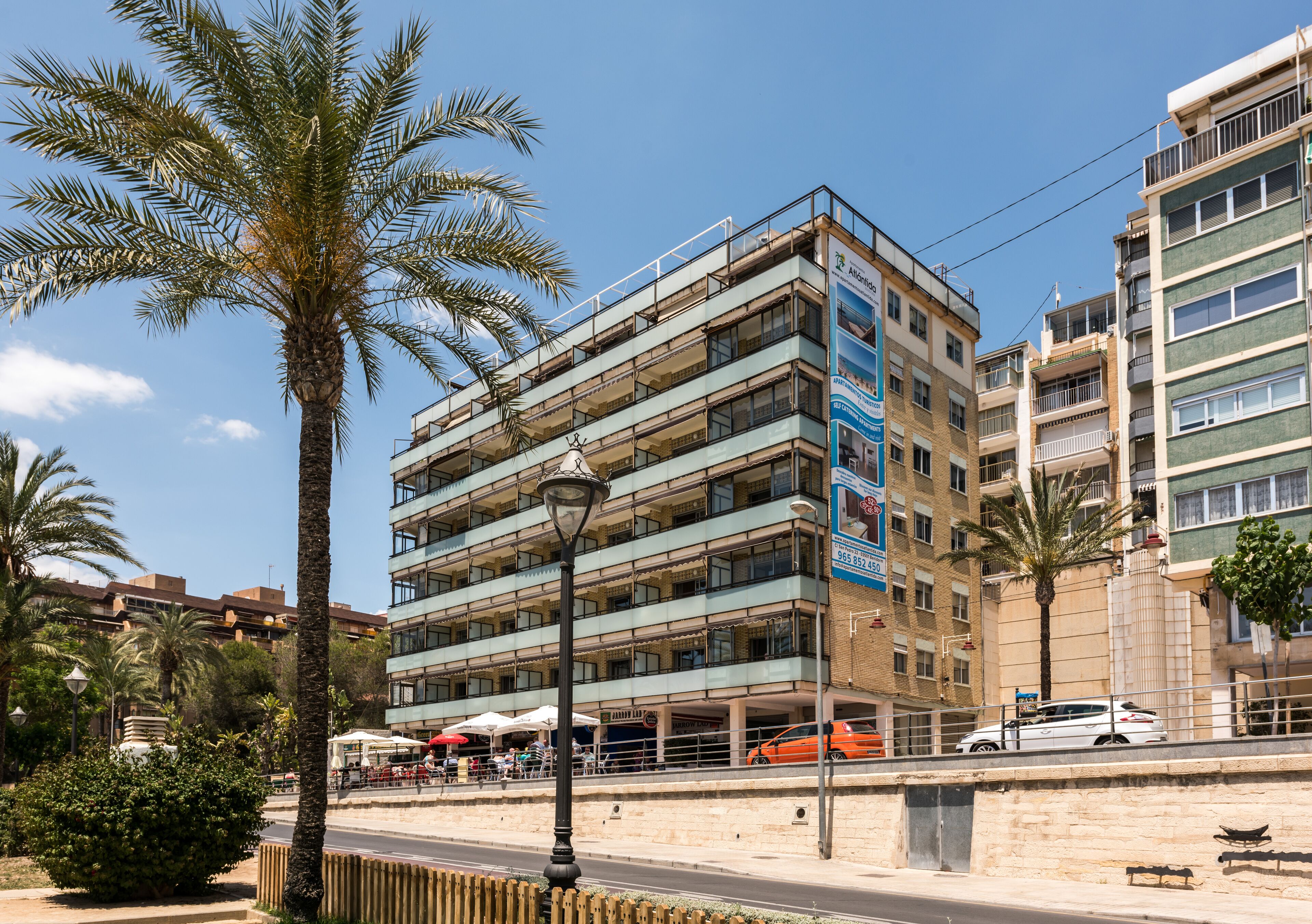 A multi-story building with balconies, a palm tree, and a street with parked cars.